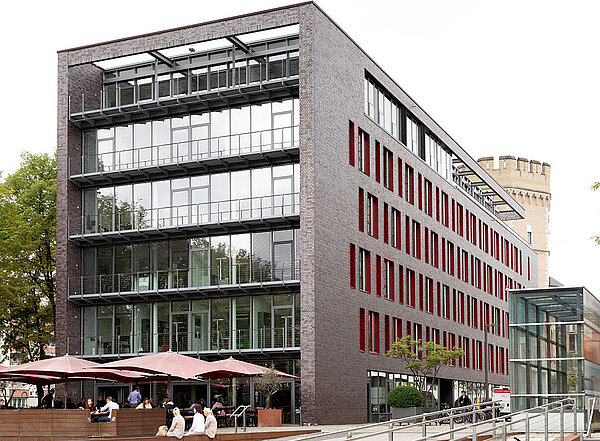 Ein modernes Gebäude mit großen Glasfronten und roten Fenstern. Vor dem Gebäude befinden sich ein rampenförmiger Zugang und ein Außenbereich mit Sonnenschirmen, in dem Menschen sitzen. Zur rechten Seite steht ein Fahrrad.
