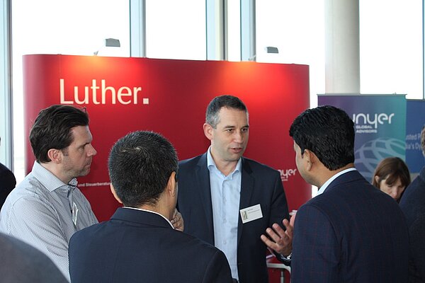 A group of four men engage in a discussion at a networking event. One man, gesturing as he speaks, stands out among the others, who are actively listening. The background features a large display with the text "Luther."