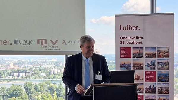 A man in a suit stands at a podium, speaking at a law firm event. Behind him is a banner for "Luther," showcasing their global locations with images of various cities. The view through the window shows a landscape with trees and buildings.