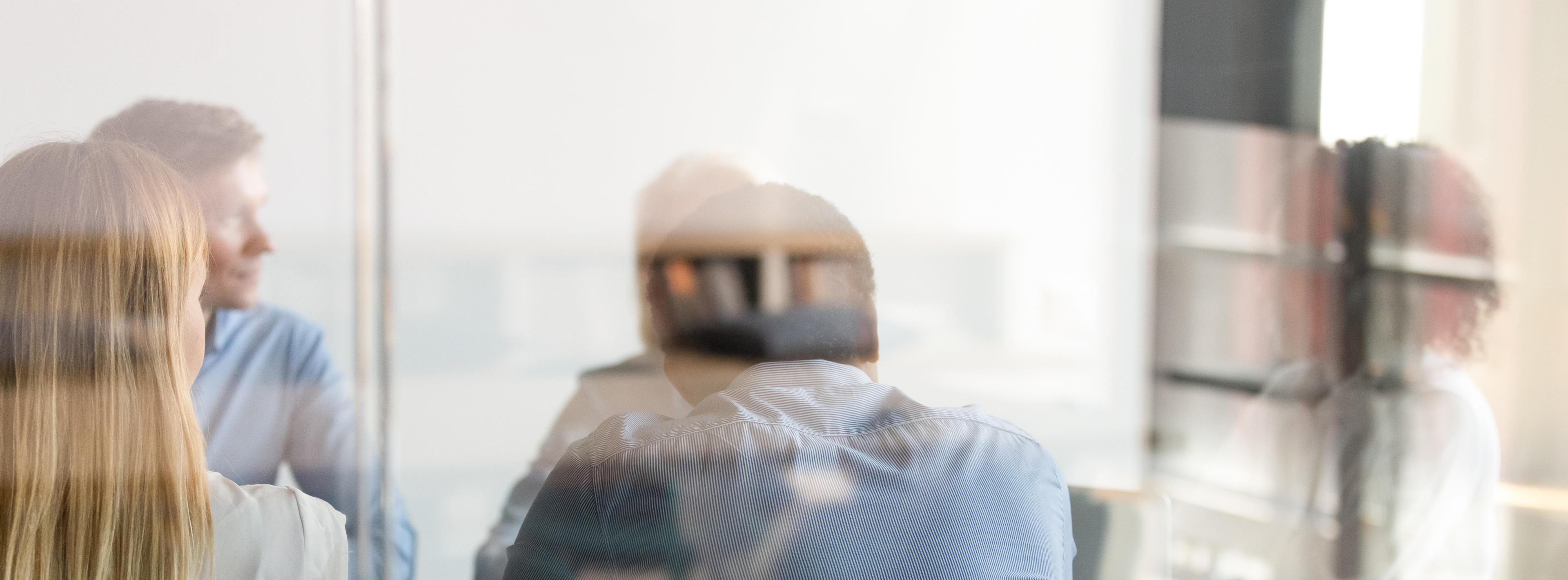 A group of professionals sit in a meeting room, engaging in discussion. Their backs are turned towards the viewer, with soft reflections visible on the glass wall. The atmosphere conveys collaboration and focused dialogue.
