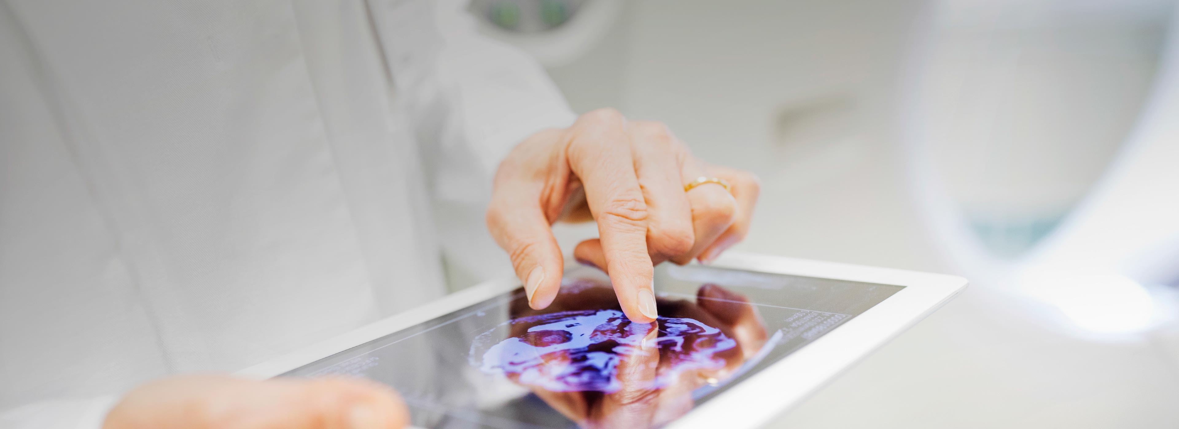 A close-up image of a hand pointing at a digital tablet displaying a medical scan, possibly a brain scan. The background is blurred, focusing attention on the screen, which shows intricate patterns in varying shades of purple and blue.