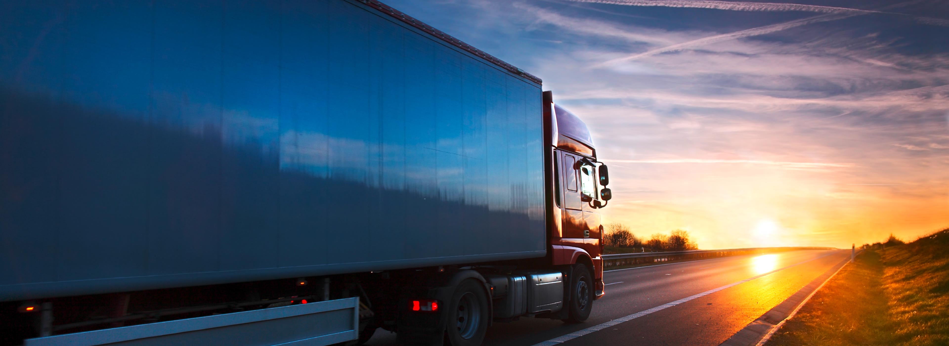 A large truck drives along a highway at sunset. The sky is filled with warm hues of orange and blue. The road stretches ahead, illuminated by the fading sunlight, creating a serene atmosphere.