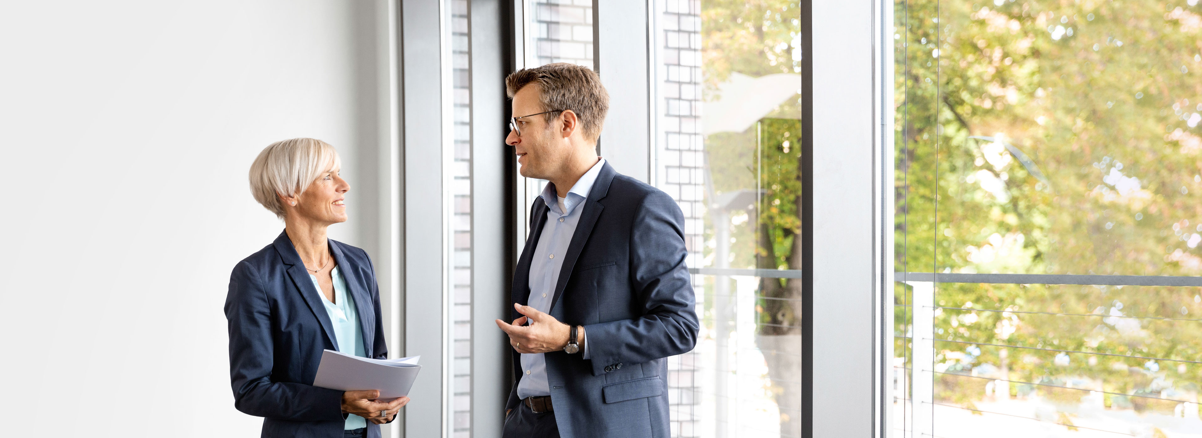 Two professionals are engaged in conversation inside a modern office space, with large windows showing greenery outside. The woman holds a stack of papers, and both are dressed in business attire. They appear to be discussing work-related topics.
