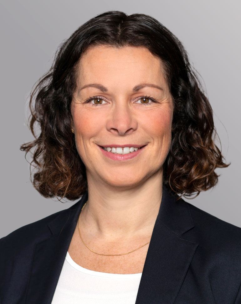 A woman with shoulder-length curly hair is smiling. She is wearing a dark blazer over a white top and a delicate gold necklace. The background is a soft gray gradient. Her expression is friendly and approachable.