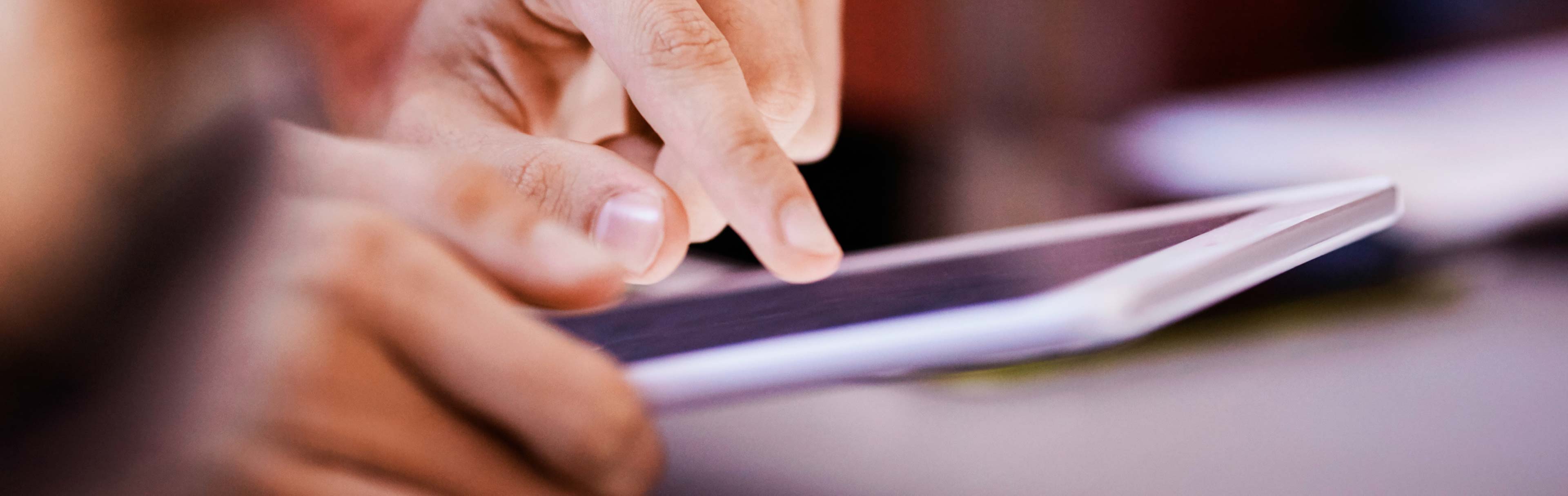 A close-up of a person's hands interacting with a tablet screen, highlighting fingers tapping the device. The background is softly blurred, emphasizing the focus on the hands and the technology.