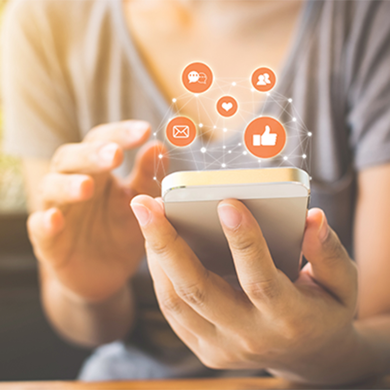 A person is holding a smartphone, interacting with various social media icons above the device. Icons include chat, heart, thumbs up, and email symbols, representing communication and online engagement. The background is softly lit, suggesting a warm atmosphere.