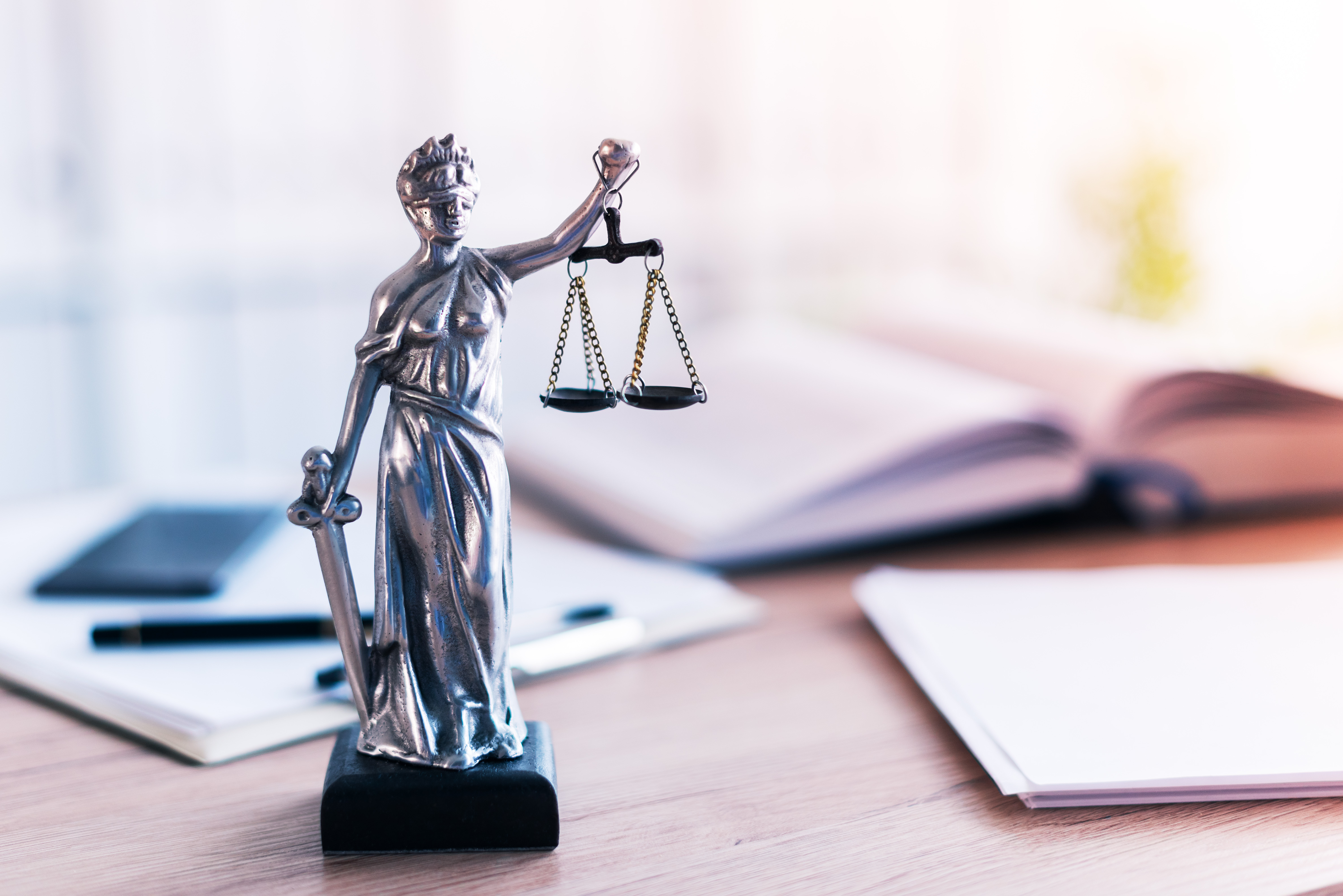 A silver statue of Lady Justice holding scales in one hand and a sword in the other, symbolizing law and fairness. The background includes blurred books and stationery items, indicating a legal or scholarly setting.