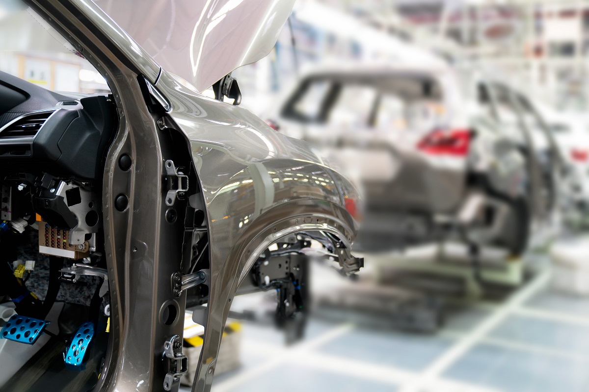 The image shows the interior of a car in a factory setting, focusing on the open door revealing components like the dashboard and pedals. In the background, other vehicles are seen in various stages of assembly. The environment appears busy and well-lit, typical of an automotive production line.