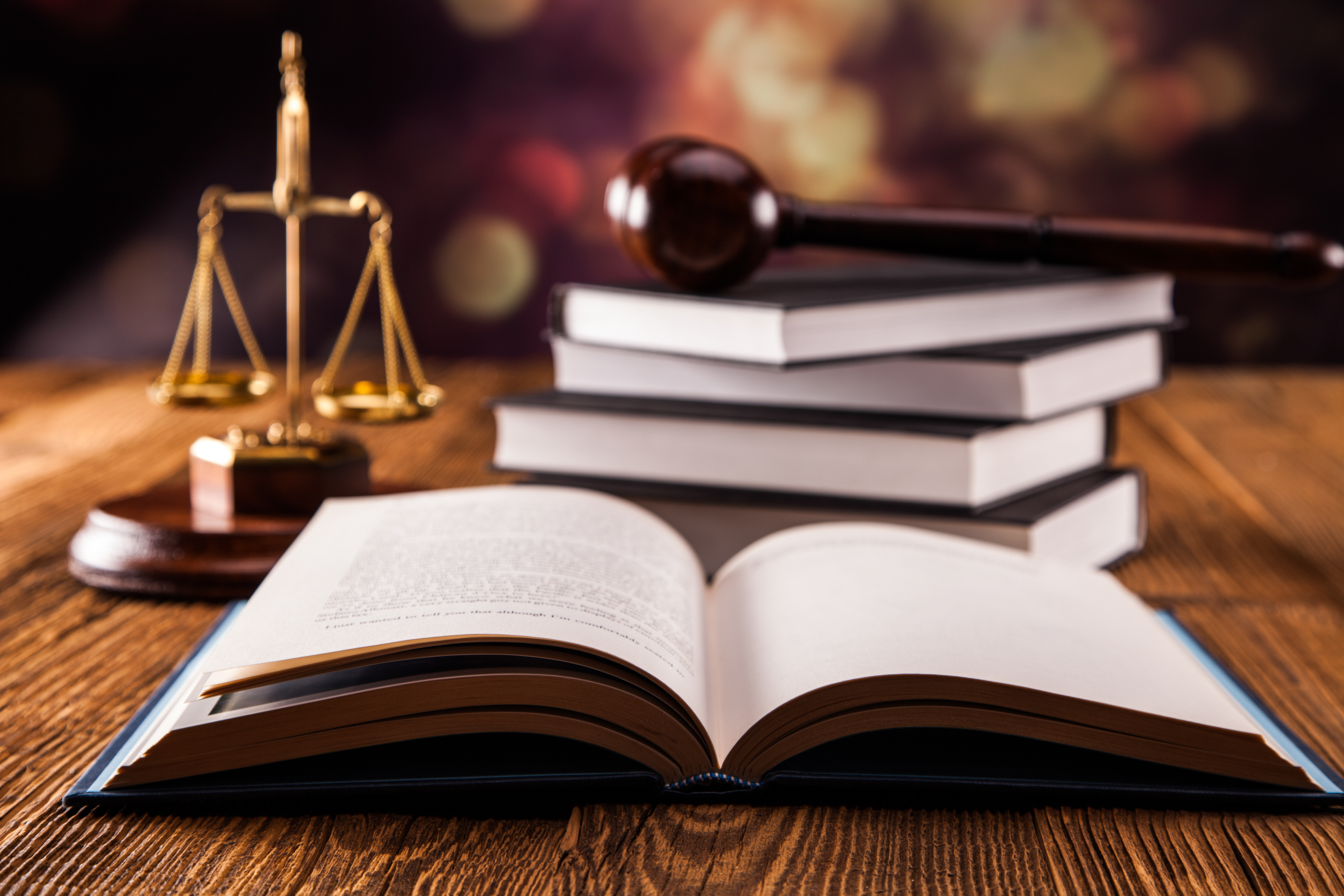 Ein offenes Buch liegt auf einem Holztablett. Im Hintergrund sind gestapelte Bücher sichtbar. Daneben steht eine goldene Justitia-Waage und ein Richterhammer. Die Szene vermittelt eine rechtliche Atmosphäre, passend zu Themen von Recht und Gesetz.