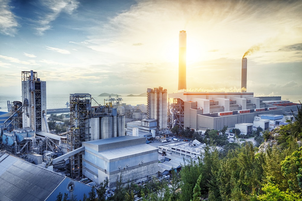 Eine Industrieanlage im Vordergrund mit großen silbernen Silos und hohen Schornsteinen, aus denen Rauch aufsteigt. Im Hintergrund ist eine Küstenlandschaft zu sehen, während die Sonne über dem Wasser aufgeht und den Himmel in warmen Farben erleuchtet.