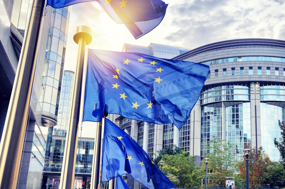 Image shows multiple European Union flags waving in front of modern glass buildings. The scene captures a sunny day, enhancing the vibrant colors of the flags. The setting reflects an official or governmental atmosphere associated with the European Union.