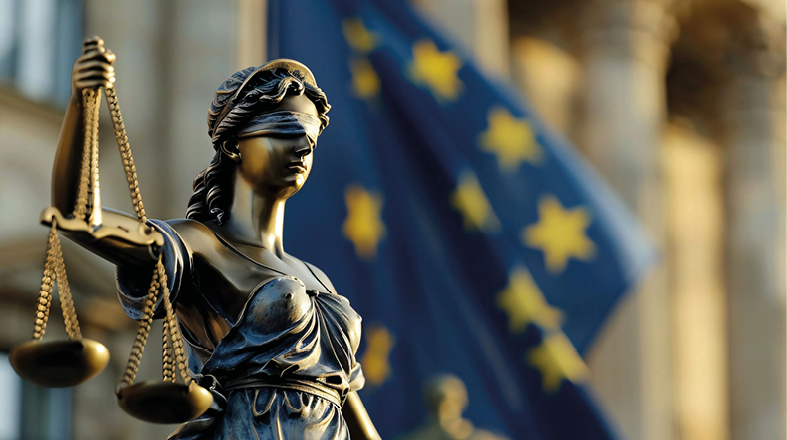 A bronze statue of Lady Justice stands holding scales, symbolizing impartiality and fairness. She is blindfolded, representing objectivity in the legal system. In the background, an out-of-focus flag with stars is visible, suggesting a European context.