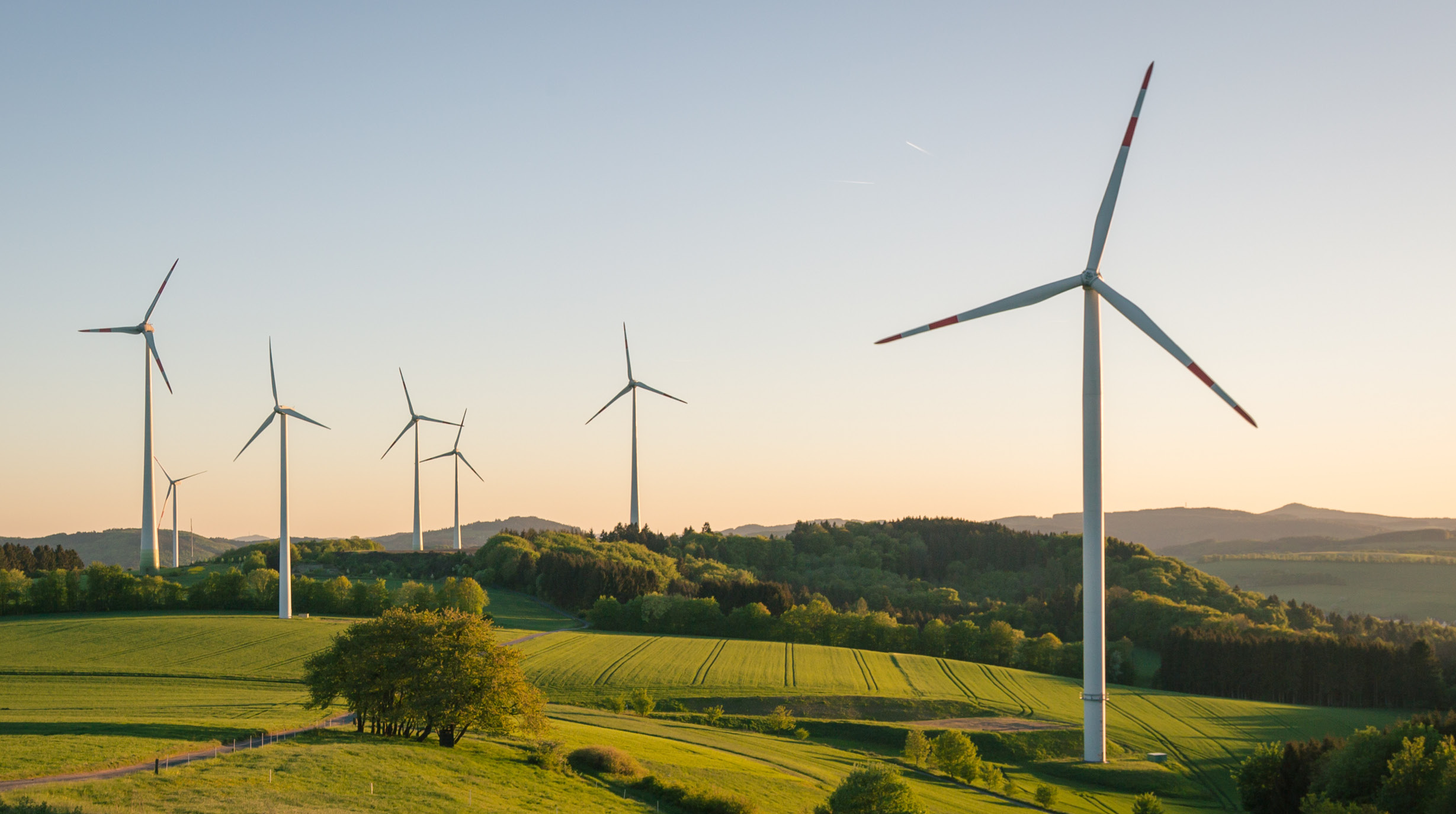 Sechs Windkraftanlagen stehen auf einem grünen Hügel in einer ländlichen Umgebung. Die Rotorblätter sind in Bewegung, während die Sonne am Horizont aufgeht. Im Hintergrund sind sanfte Hügel und Bäume sichtbar.