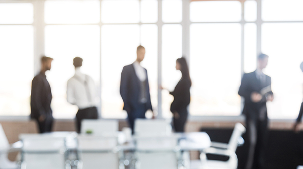 A group of professionals in business attire engage in conversation in a bright, modern office space. The focus is on their interactions against large windows that let in natural light, suggesting a collaborative work environment.