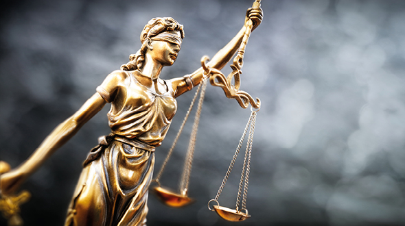 A bronze statue of Lady Justice, blindfolded, holds a balanced scale in one hand and a sword in the other. She represents fairness and the impartiality of the law, symbolizing justice and truth. The background is a blurred gray, emphasizing the figure.