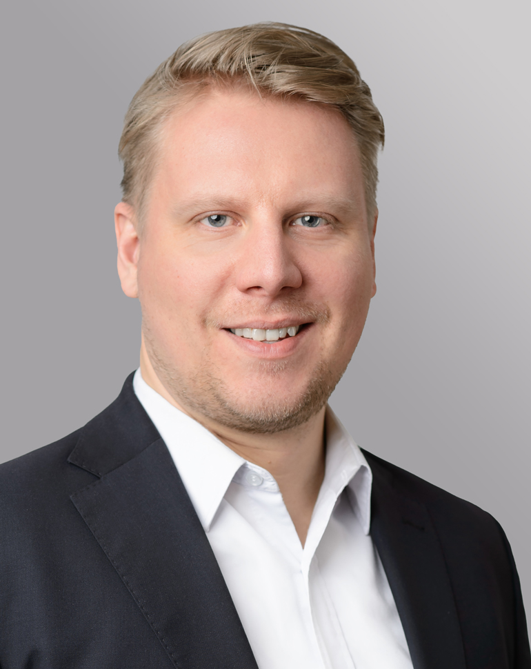 Porträt eines lächelnden Mannes mit blonden Haaren, der einen grauen Anzug und eine gestreifte Krawatte trägt. Der Hintergrund ist neutral und grau, was den Fokus auf das Gesicht des Mannes legt.