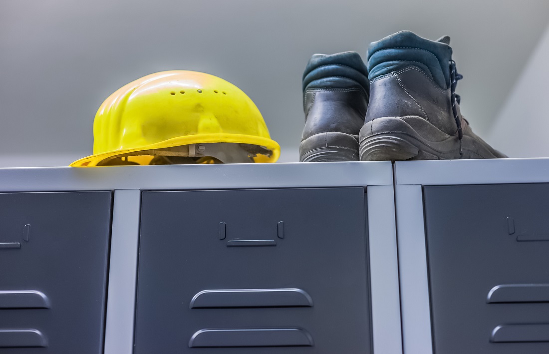 A bright yellow hard hat sits atop a row of gray lockers, with a pair of sturdy work boots positioned beside it. The scene indicates a work environment focused on safety and construction.