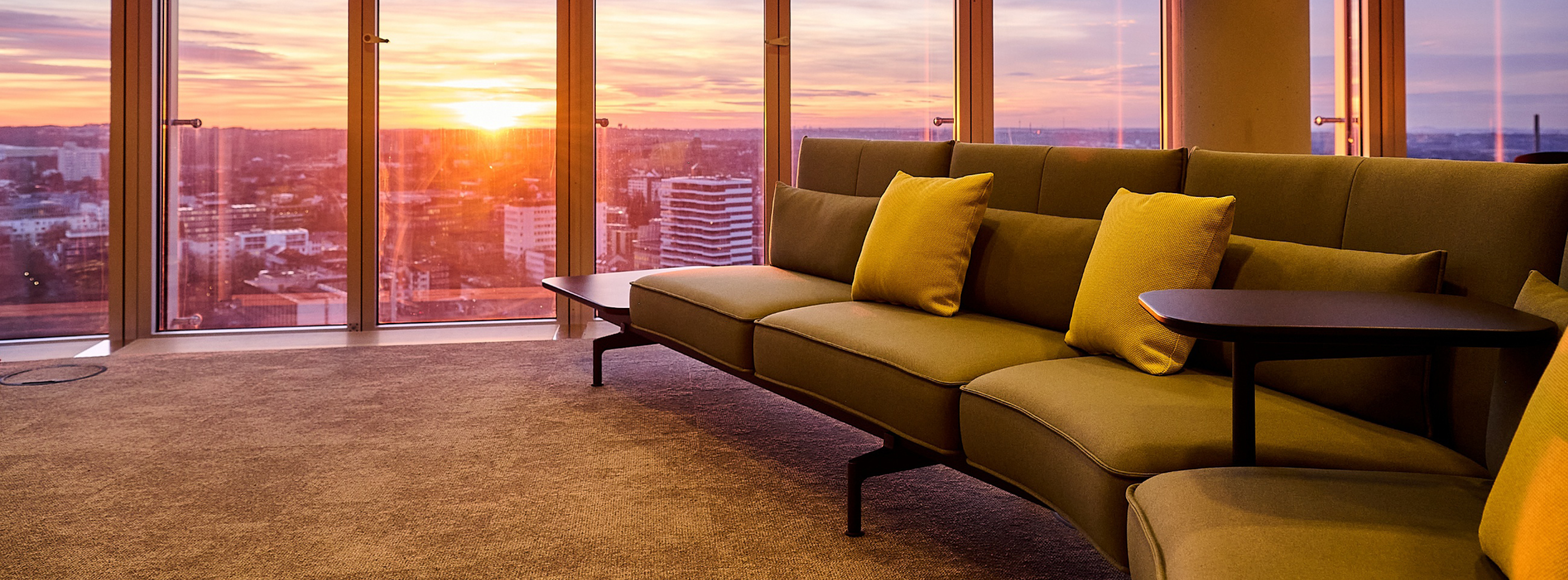 Ein modernes Wohnzimmer mit einer beigen Couch und gelben Kissen. Große Fenster bieten einen spektakulären Blick auf einen Sonnenuntergang über der Stadt. Der Raum hat einen eleganten, minimalistischen Stil und ist mit einem weichen Teppich ausgelegt.