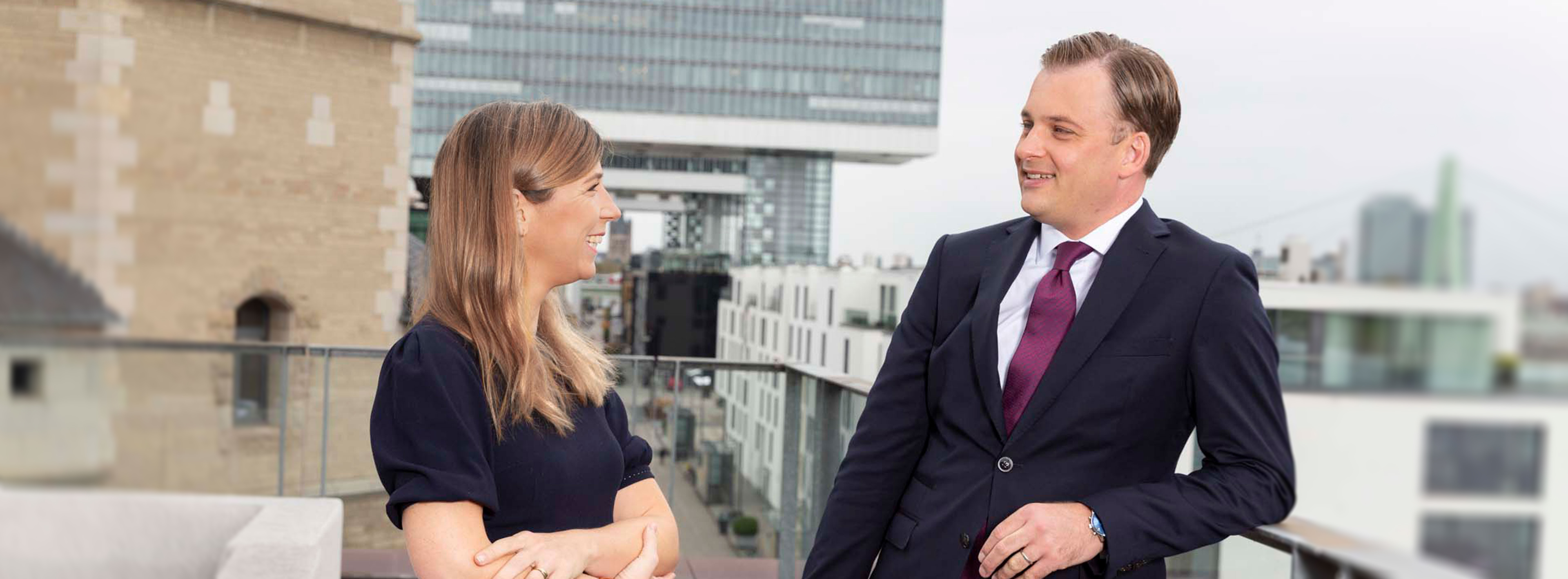 Two professionals, a man and a woman, are smiling and engaged in conversation on a balcony with a city skyline in the background. The woman is wearing a dark dress, while the man is dressed in a suit with a tie.