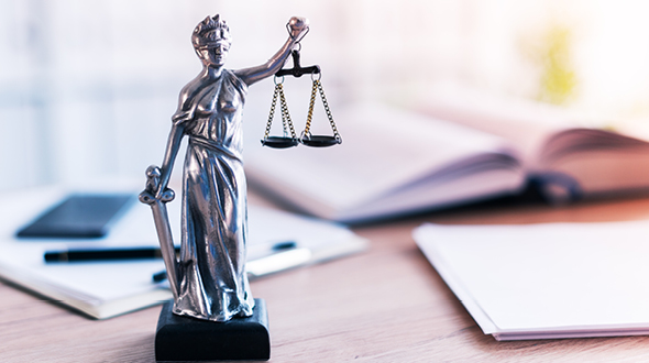 A silver figurine of Lady Justice stands on a desk, holding balanced scales in one hand and a sword in the other, symbolizing law and fairness. In the background, there are blurred books and notepads, suggesting a legal or scholarly setting.
