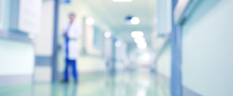 Unscharfe Aufnahme eines Krankenhausflurs mit grünlichen Wänden und Beleuchtung. Im Hintergrund ist eine verschwommene Person in einem weißen Kittel zu sehen. Der Flur wirkt ruhig und ordentlich, mit reflektierendem Boden.