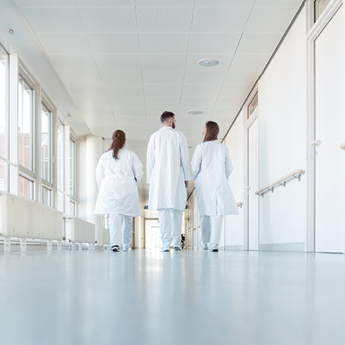 Three medical professionals in white lab coats walk together down a well-lit hospital corridor. Their backs are to the viewer as they proceed along a smooth floor, flanked by large windows that allow natural light into the space.