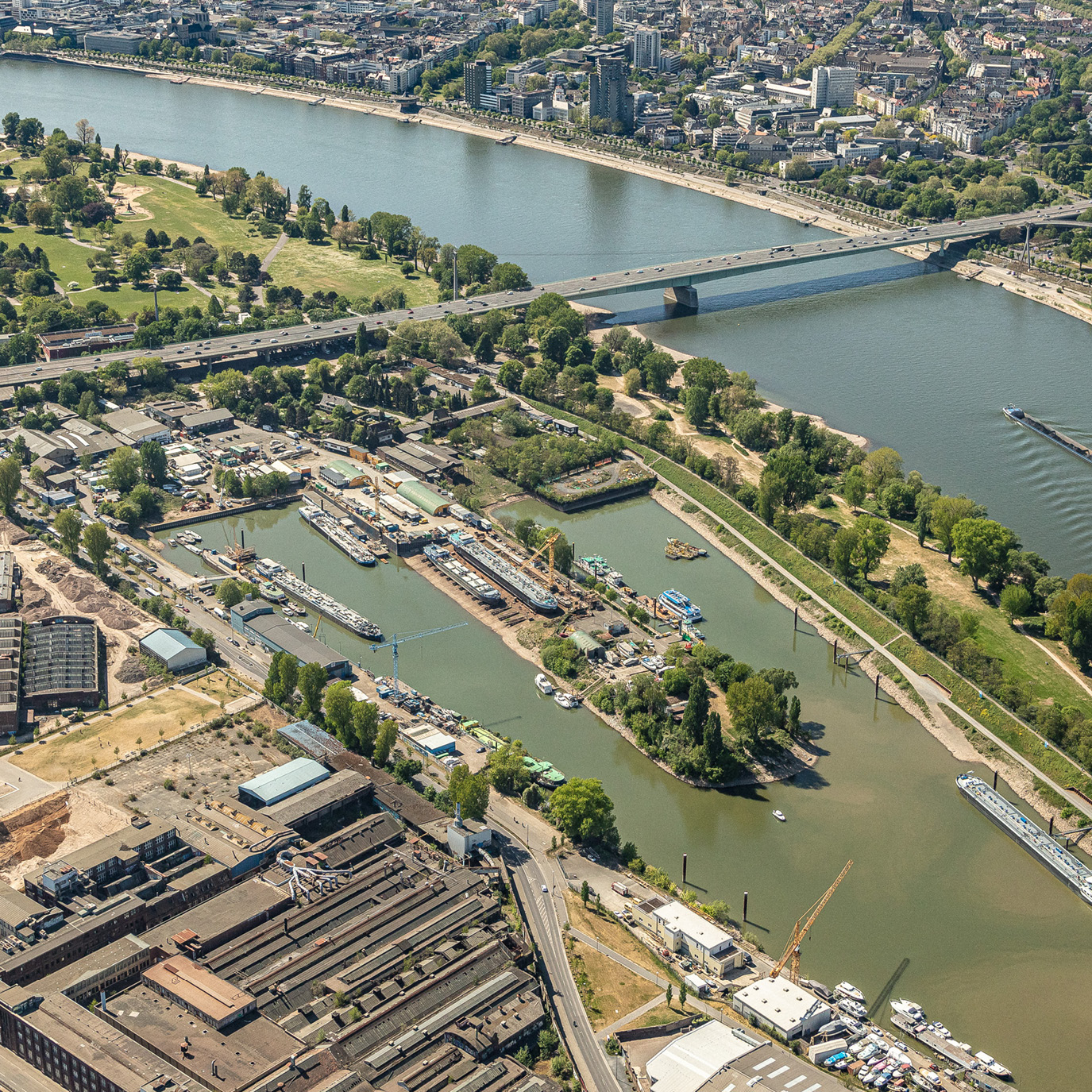 Luftaufnahme eines Flusses, umgeben von Grünflächen und städtischen Gebäuden. An den Ufern sind mehrere Schiffe und Boote zu sehen, während eine Brücke den Fluss überspannt. Im Vordergrund sind Industrieanlagen erkennbar.