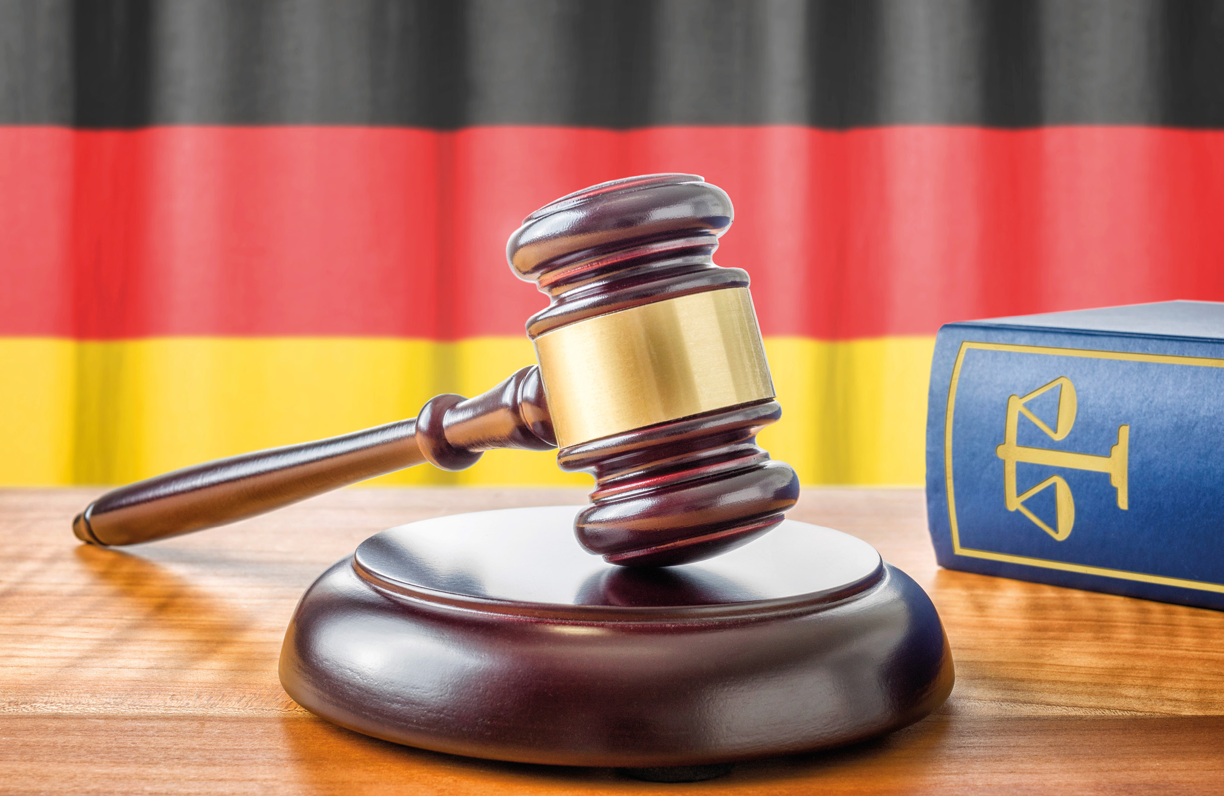 Ein Richterhammer liegt auf einem Holzblock, daneben ein blaues Buch mit einer Waage als Symbol für Gerechtigkeit. Im Hintergrund ist eine Gardine mit den Farben der deutschen Flagge (Schwarz, Rot, Gold) sichtbar.