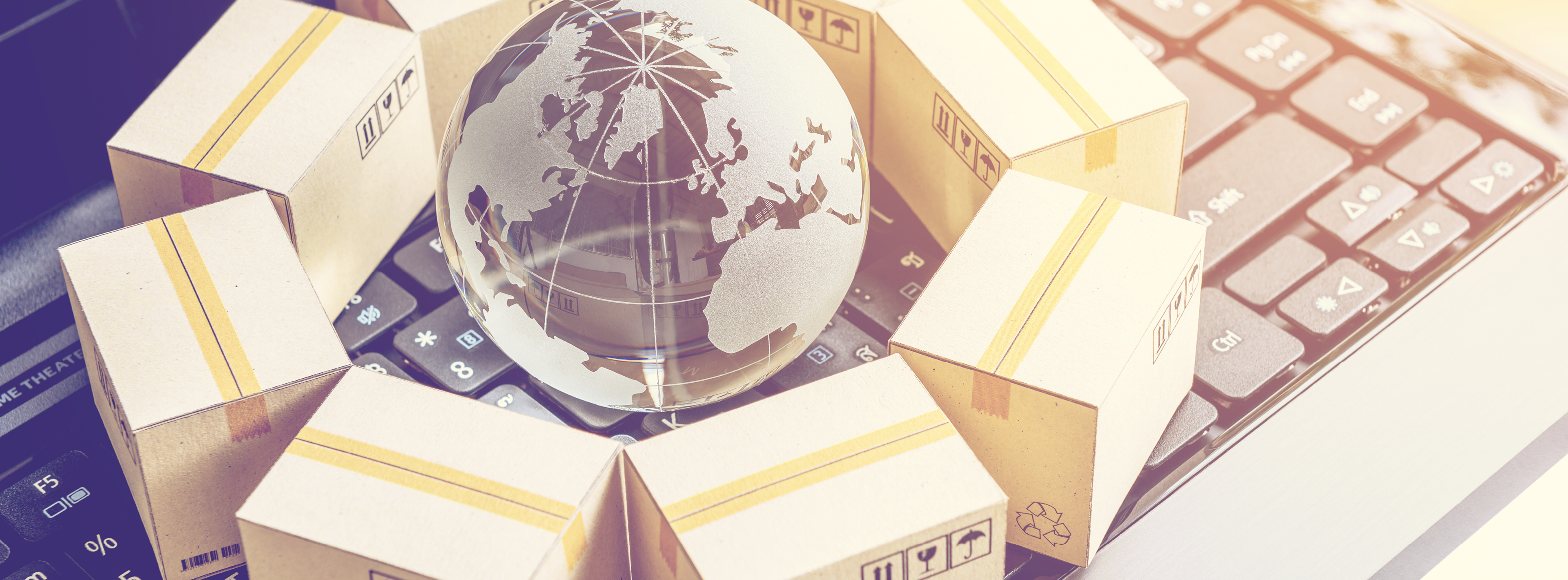 Eine gläserne Weltkugel steht auf einer Computertastatur, umgeben von mehreren geschlossenen Kartons. Die Anordnung symbolisiert den internationalen Versand und die Vernetzung im digitalen Handel.