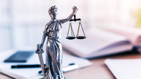A silver statue of Lady Justice stands holding scales in one hand and a sword in the other. She is draped in a flowing robe and wears a blindfold, symbolizing impartiality in the legal system. The background features blurred office materials, suggesting a legal setting.