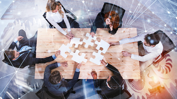 A diverse group of six professionals collaborates around a wooden table, holding pieces of a large puzzle. They appear engaged in discussion, symbolizing teamwork and problem-solving. The background suggests a digital or innovative environment.