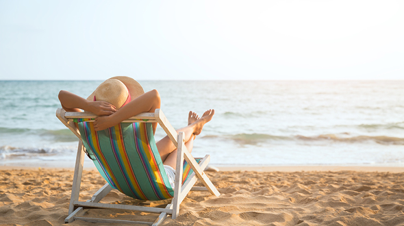 Eine Person entspannt in einem Liegestuhl am Strand, mit einem Sonnenhut auf dem Kopf. Die Wellen des Meeres sind im Hintergrund sichtbar, während die Sonne am Horizont untergeht. Der Strand ist mit feinem Sand bedeckt.