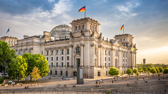 Das Bild zeigt das Reichstagsgebäude in Berlin, ein historisches Bauwerk mit einer markanten Glaskuppel. Im Vordergrund sind Bäume und Bürger zu sehen, die den Platz um das Gebäude nutzen. Deutsche Flaggen wehen am Gebäude in der Abendbeleuchtung.