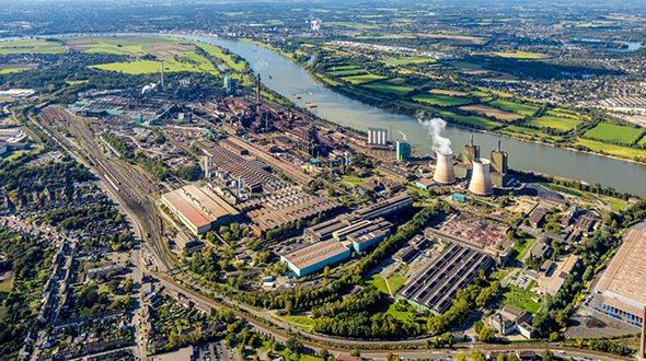 Luftaufnahme eines großen industriellen Areals mit Fabrikgebäuden, Kühltürmen und einem Fluss im Hintergrund. Umgeben von grünen Feldern und Wohngebieten, zeigt die Szene einen modernen industriellen Standort.