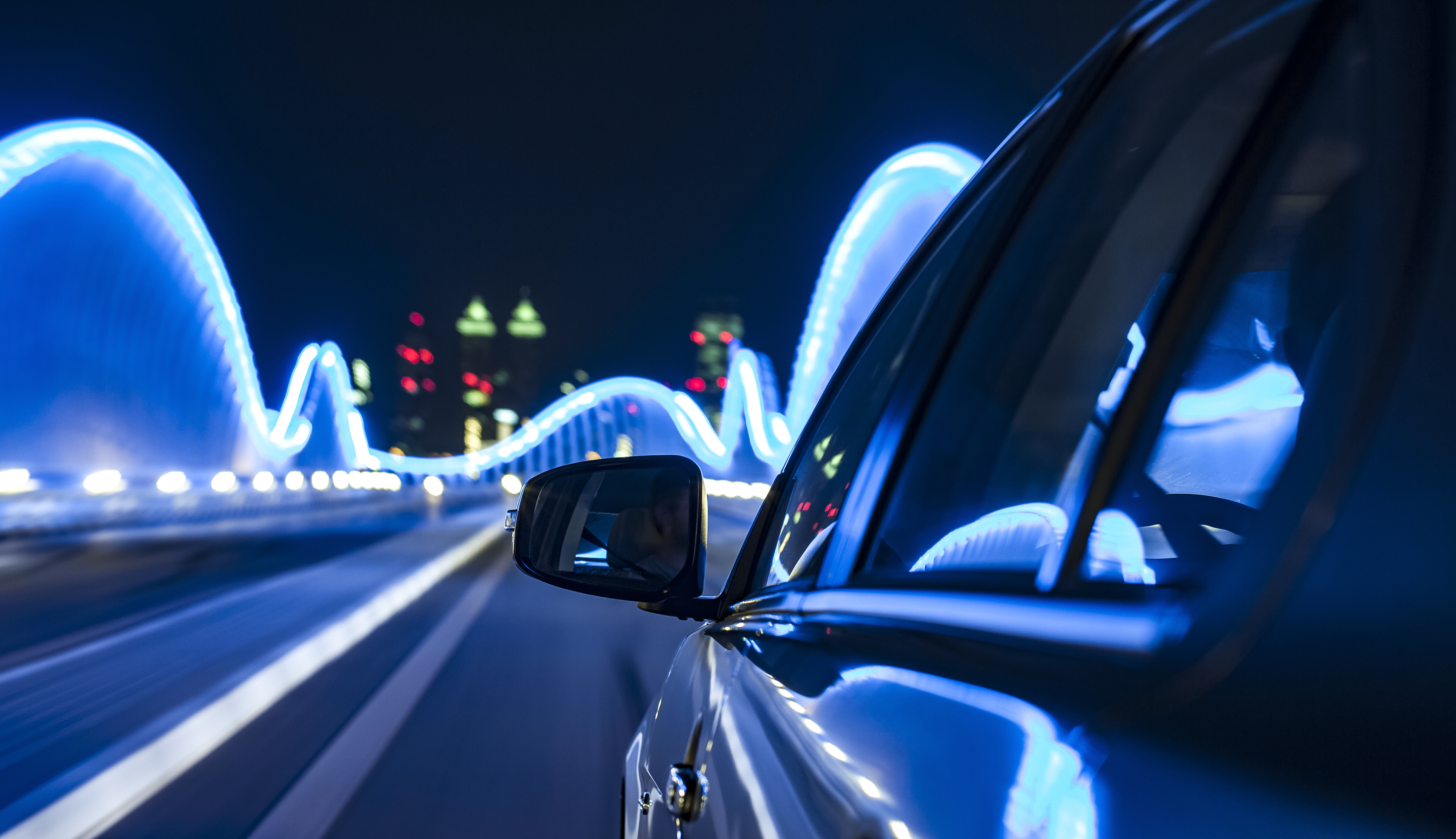 A close-up view from inside a car, showing the side mirror and street lights, as the vehicle drives through a city at night. Bright blue neon lights create a dynamic pattern around the bridge in the background, suggesting a sense of speed and movement.