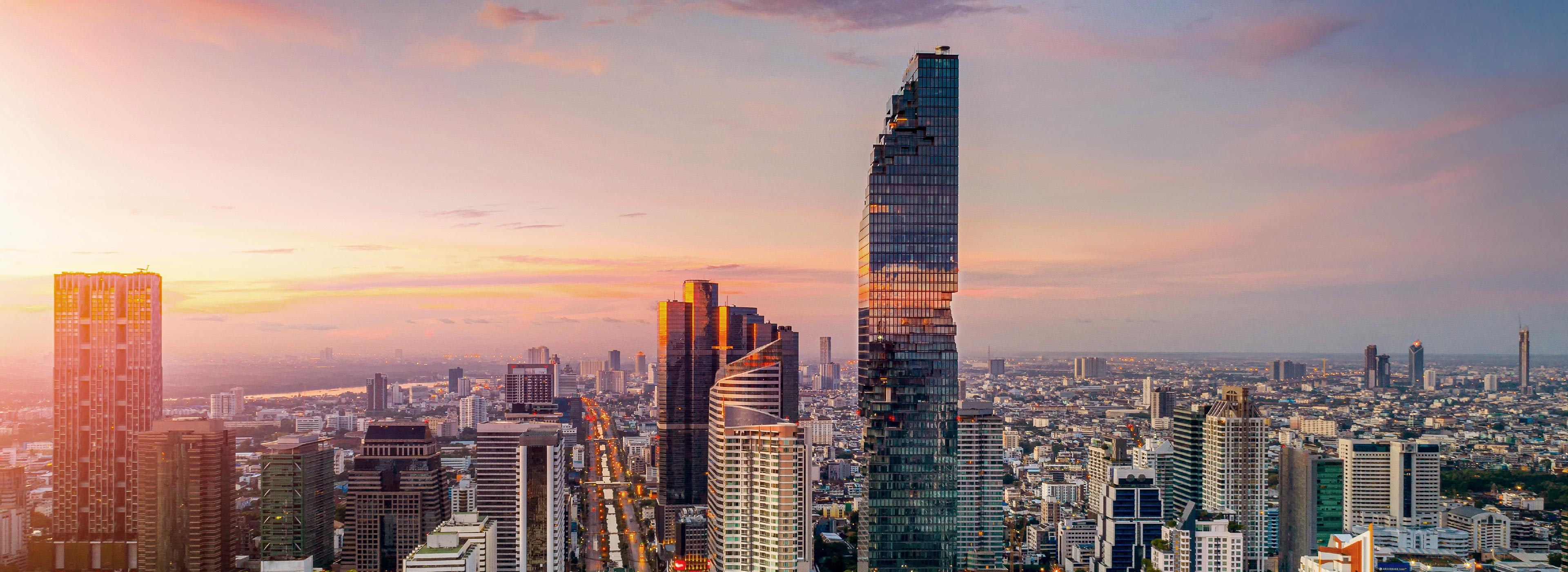 A panoramic view of a vibrant urban skyline at sunset, showcasing a mix of modern skyscrapers. The tallest building, characterized by a unique architectural design, stands prominently against a colorful sky with soft clouds, while the city's streets and buildings stretch into the distance.
