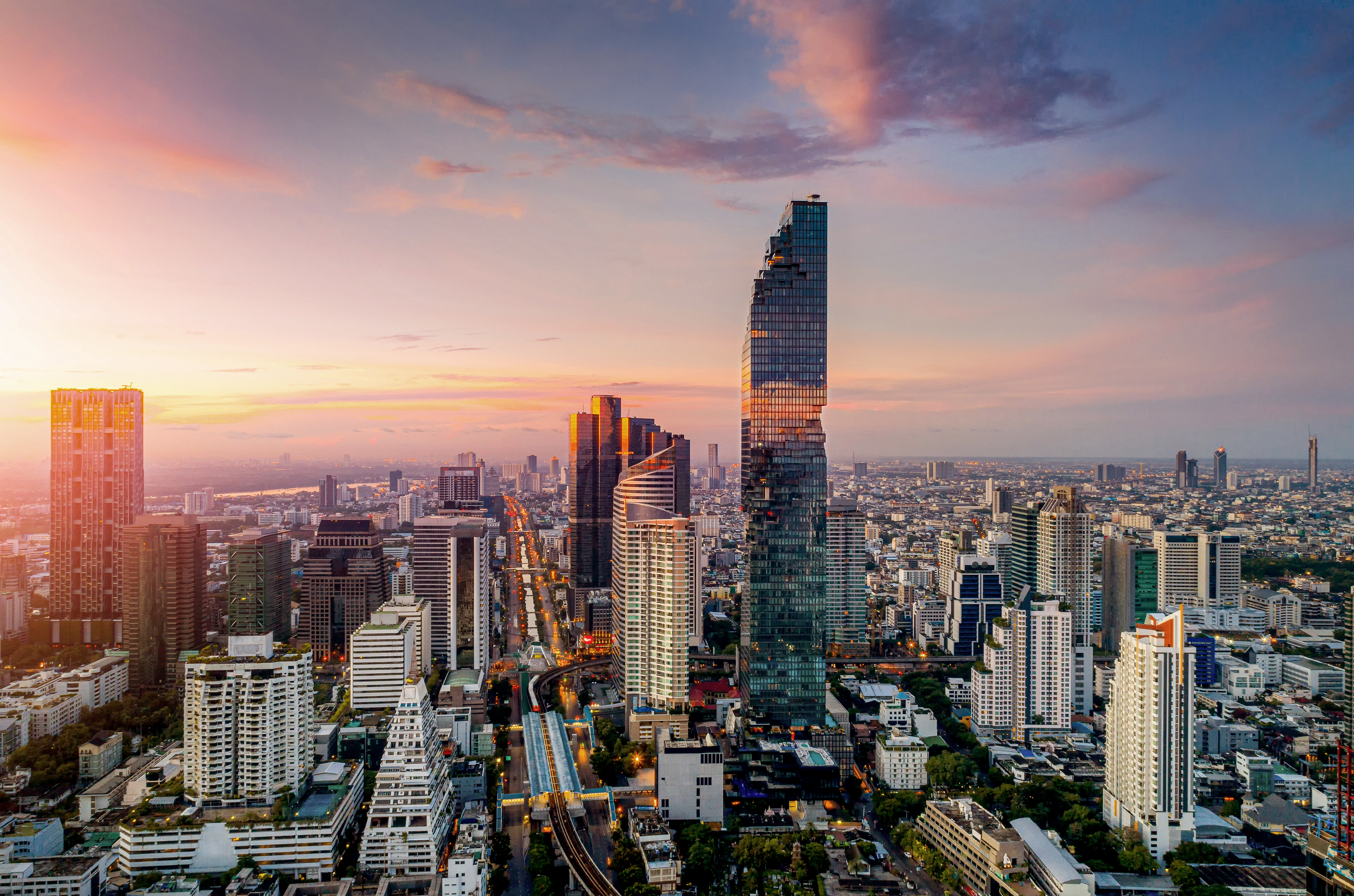 A city skyline at sunset shows a blend of modern skyscrapers and buildings, reflecting warm orange and gold hues. The streets below are lined with structures of various architectural styles, creating a vibrant urban landscape. The overall atmosphere is lively and dynamic.