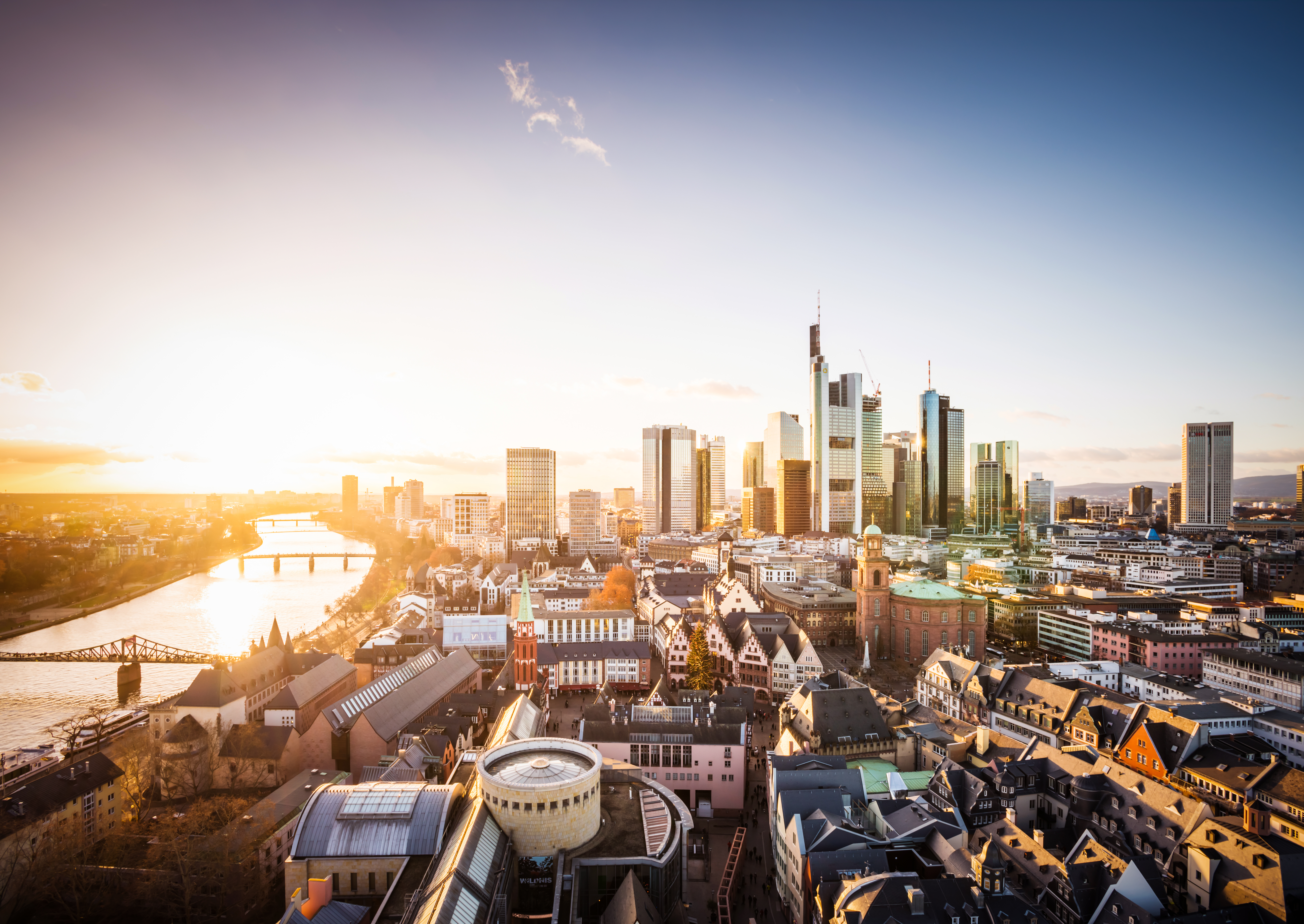 Luftaufnahme der Frankfurter Skyline bei Sonnenuntergang. Hochmoderne Wolkenkratzer illustrieren die Architektur der Stadt, während die ruhigen Wasser des Mains im Vordergrund reflektieren. Historische Gebäude sind im Stadtbild integriert, was einen Kontrast zwischen Alt und Neu zeigt.