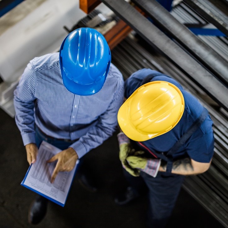 Two individuals in safety helmets—one blue and one yellow—are discussing a document. They are in a workshop environment with metal materials in the background. The person in the blue helmet is pointing at the document while the other listens attentively.