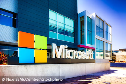 Das Bild zeigt das Außenansicht eines modernen Gebäudes mit dem großen Microsoft-Logo im Vordergrund. Das Logo besteht aus bunten Quadraten in Orange, Grün, Blau und Rot, neben dem Schriftzug "Microsoft" in großer, weißer Schrift.