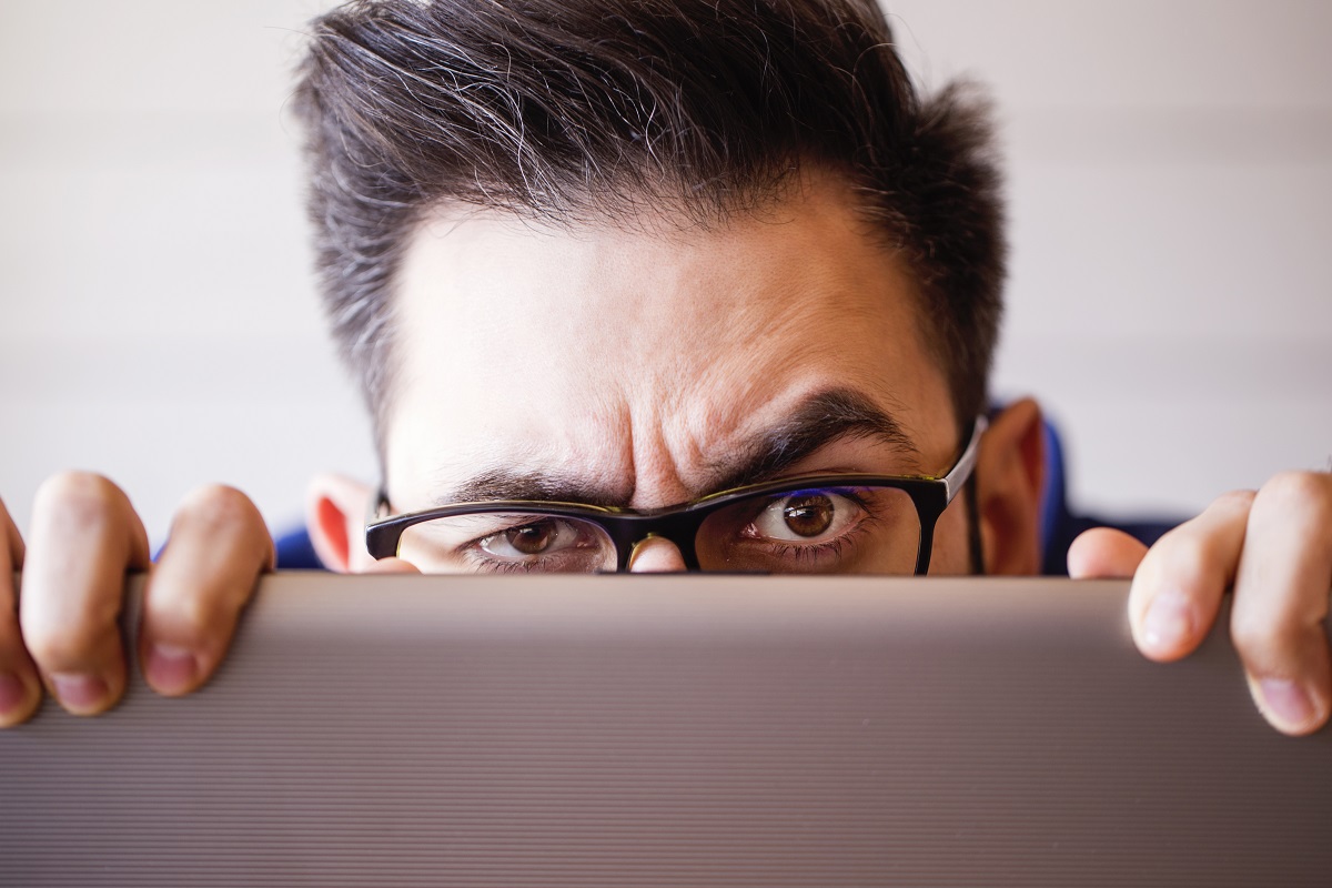 A man with glasses is peering over a laptop, displaying a concerned expression. His furrowed eyebrows and intense gaze suggest curiosity or apprehension about what he is seeing on the screen.