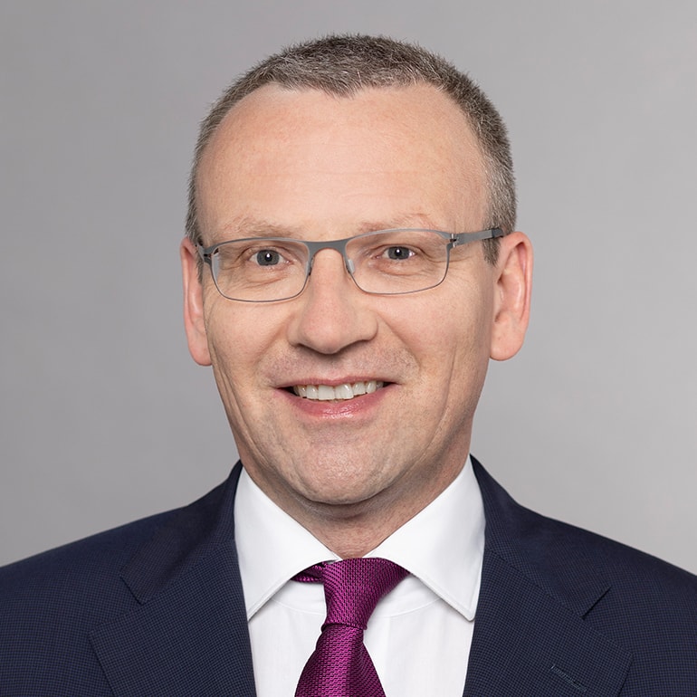 A middle-aged man with short, light hair and glasses smiles at the camera. He is wearing a dark suit and a white shirt with a purple tie. The background is a subtle gray, providing a neutral backdrop for his professional appearance.