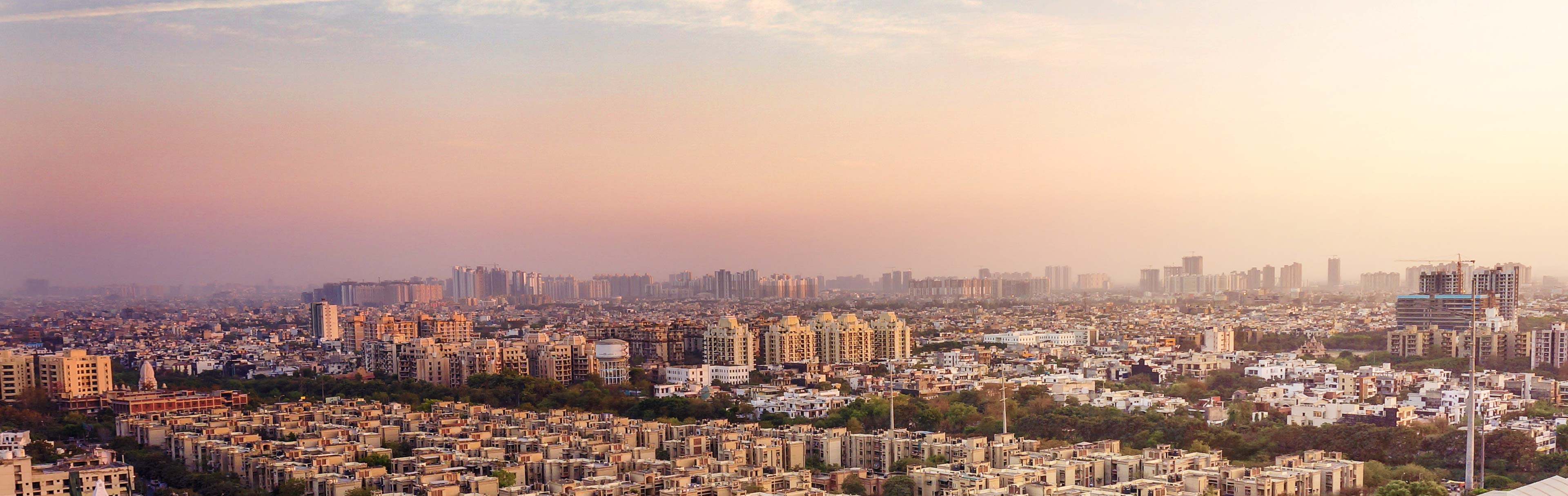 A panoramic view of a sprawling urban landscape at dusk, with a mix of low-rise and high-rise buildings under a soft, pastel sky. The city is densely populated, showing a blend of residential and commercial structures stretching toward the horizon.