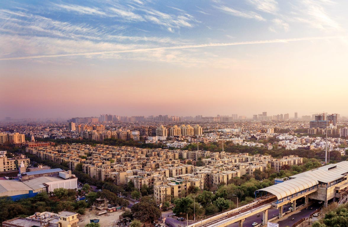 A panoramic view of a sprawling urban landscape at sunset. The foreground features clusters of buildings with trees, while the background showcases a mix of low and high-rise structures against a colorful sky, creating a sense of depth and urban development.