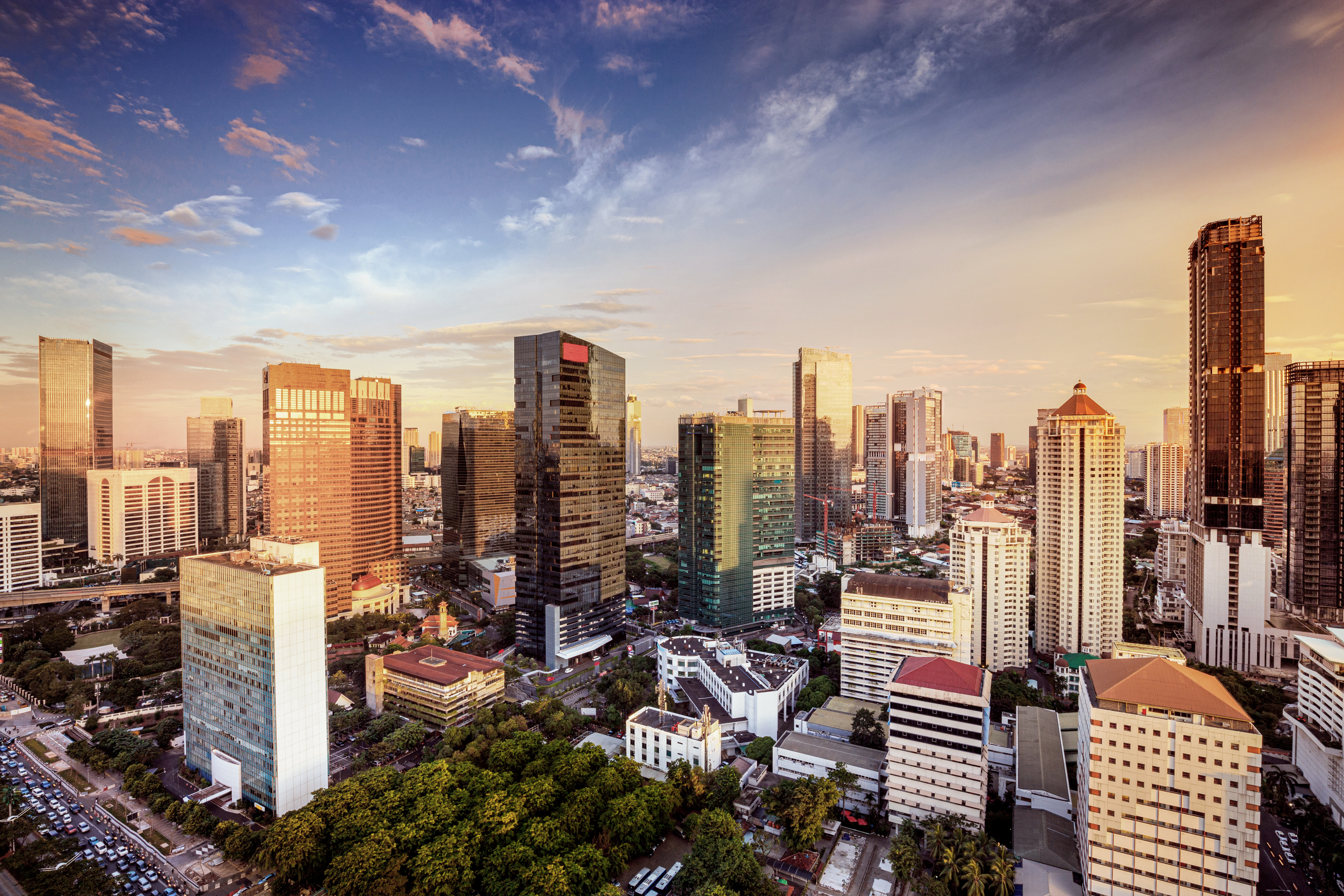 A panoramic view of a bustling city skyline during sunset. Tall skyscrapers dominate the scene, showcasing modern architecture with reflective glass surfaces. The city is partially surrounded by greenery, hinting at a vibrant urban life beneath the colorful sky.
