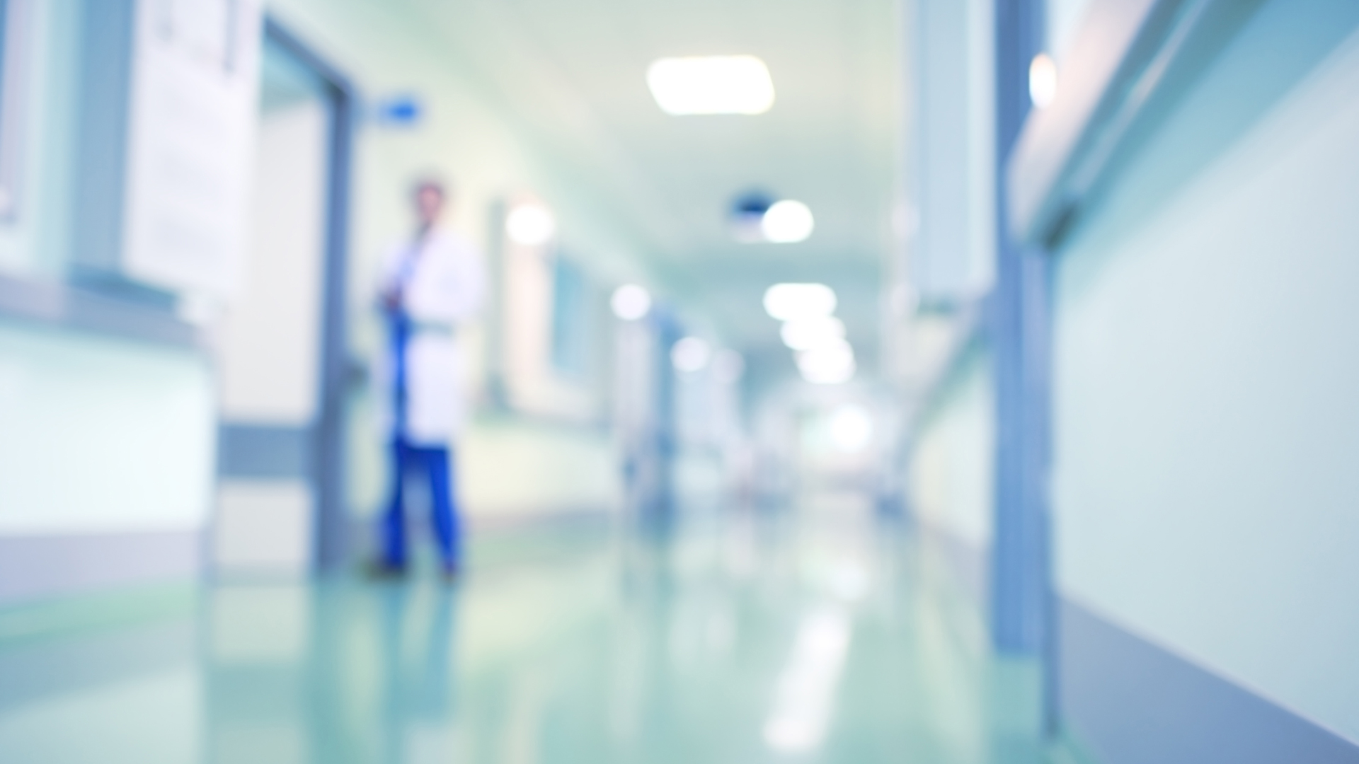 Ein unscharfes Bild eines Krankenhausflurs. Im Hintergrund steht eine Person in einem weißen Kittel. Der Flur wirkt hell erleuchtet und zeigt einige Türen und Fenster. Der Fokus liegt auf der Länge des Flurs und der Präsenz des medizinischen Fachpersonals.