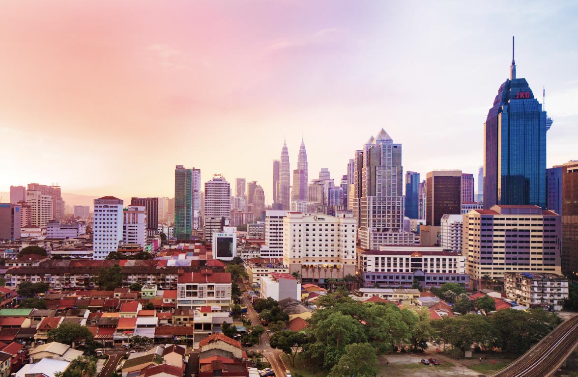 A panoramic view of a bustling urban skyline at sunset features tall skyscrapers, including two iconic twin towers. Below, a mix of modern buildings and traditional houses creates a vibrant contrast. The sky displays soft hues of pink and orange, reflecting a serene evening atmosphere.