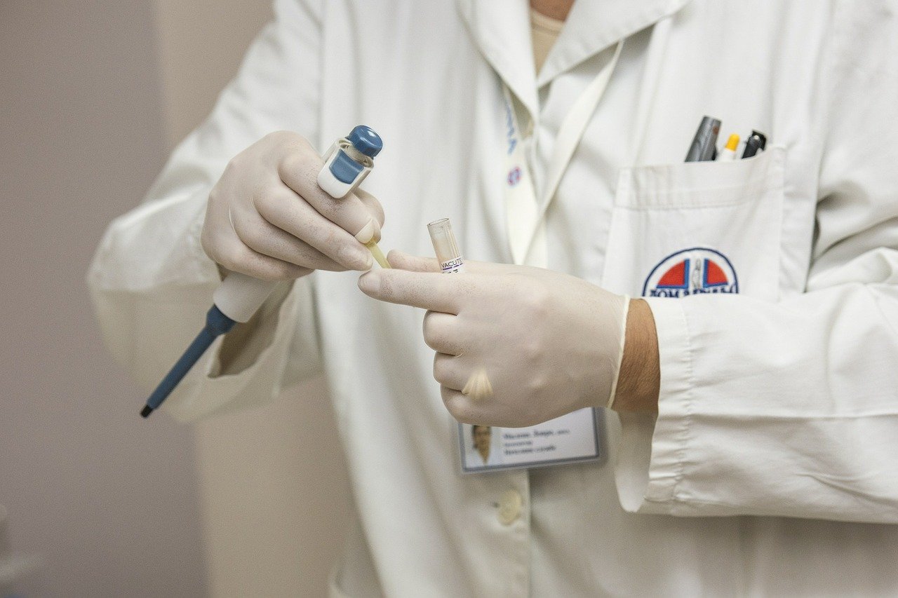 A healthcare professional wearing gloves holds a pipette in one hand and a test tube labeled "VACUT" in the other. They are preparing a sample, indicating a medical or laboratory setting focused on testing or analysis.