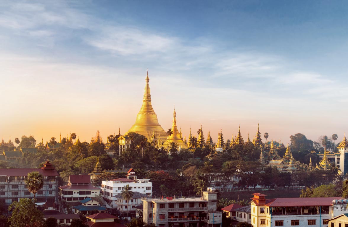A panoramic view of a golden pagoda amidst lush greenery and low-rise buildings, under a clear sky. The pagoda is surrounded by various spires and temple structures, indicating a rich cultural heritage, illuminated by the warm light of sunset.