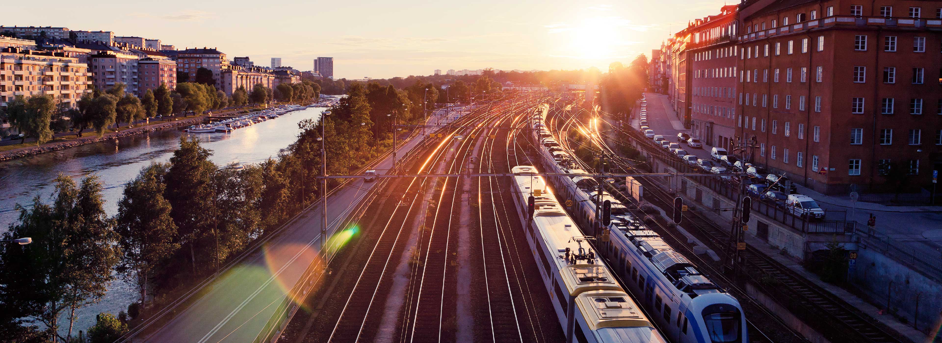 A sunset view over a railway track beside a river. The image features several trains on the tracks, surrounded by green trees and residential buildings. The warm light of the setting sun reflects off the trains and highlights the urban landscape, creating a peaceful yet vibrant scene.